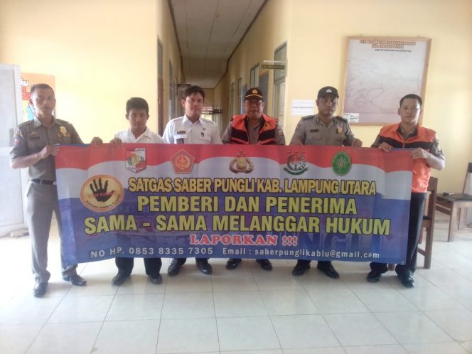 
 Foto : Humas Polres Lampura  Caption: Kasiwas Polres Lampura Ipda Sisnedi (mengenakan topi) beserta Staf kecamatan Abung Semuli saat membentangkan spanduk Satgas Saber Pungli di kecamatan setempat, foto dibidik Rabu (29/8).